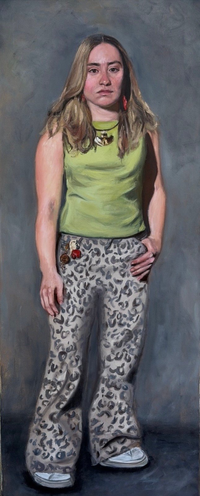 Full-length portrait of a young woman standing against a grey ground, wearing a green sleeveless top, a pendant necklace, and leopard-print flared trousers with white sneakers
