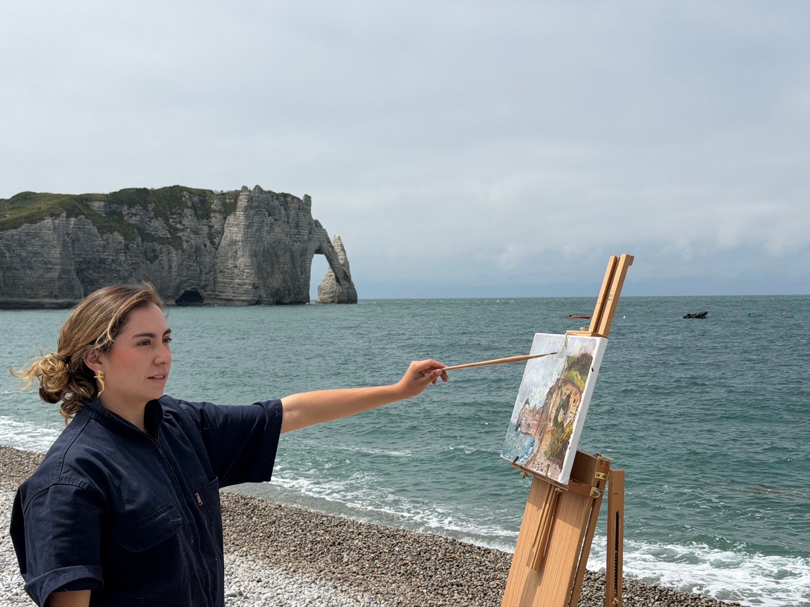 Juliana Triana painting at an easel on the pebble beach at Étretat, the chalk cliffs and the Aiguille rising from the sea behind her